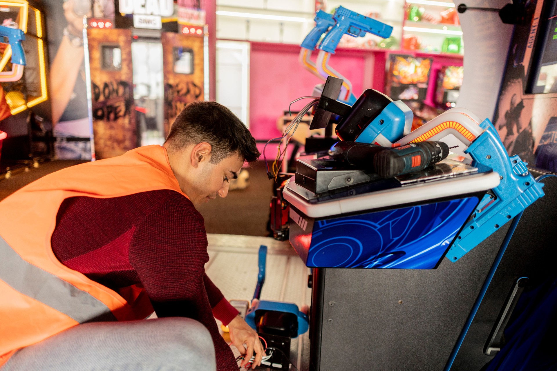 Technician Repairing Arcade Shooting Game Machine, technician in a safety vest working on repairing an arcade shooting game machine with tools in a colorful game center.